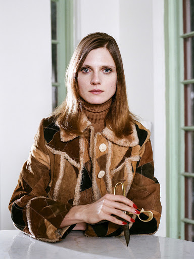 Woman with long brown hair wearing a multicolored coat stands indoors at a white table, holding a pair of gold scissors.