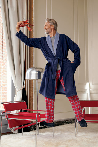 Mature adult male in robe stands on chair holding lobster indoors, wearing red plaid pants, with modern lamp and furniture.