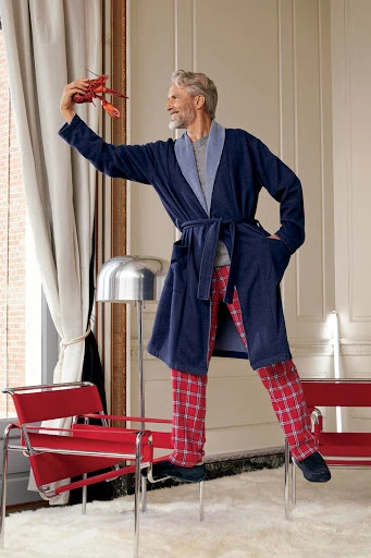 Mature adult male in robe stands on chair holding lobster indoors, wearing red plaid pants, with modern lamp and furniture.