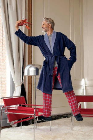 Mature adult male in robe stands on chair holding lobster indoors, wearing red plaid pants, with modern lamp and furniture.