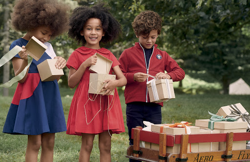 Three cheerful children in red and blue dresses and a red jacket open gift boxes outdoors, with a wagon full of presents nearby.