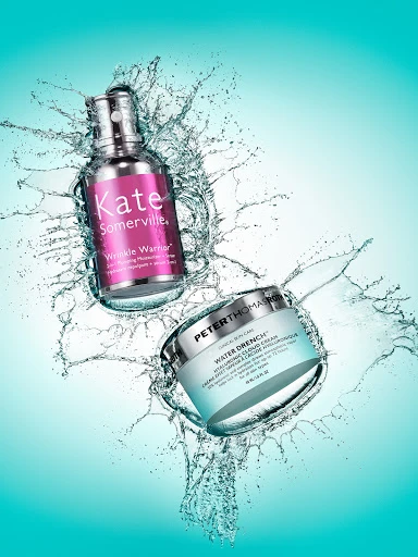 Two skincare product bottles, Kate Somerville Wrinkle Warrior and Peter Thomas Roth Water Drench, splash water against a light teal background.
