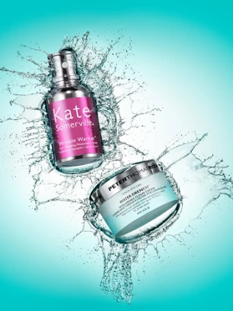 Two skincare product bottles, Kate Somerville Wrinkle Warrior and Peter Thomas Roth Water Drench, splash water against a light teal background.