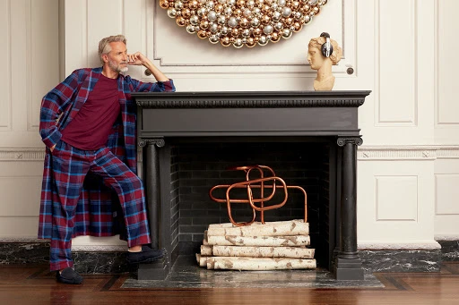 An older male model leans on a black fireplace in a blue and red plaid robe and pajamas, alongside a bust with headphones.