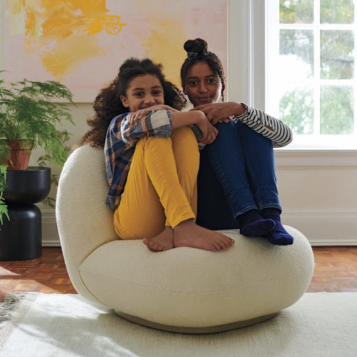 Two young girls sitting closely together on a large cream-colored, round chair indoors with natural light from a nearby window.