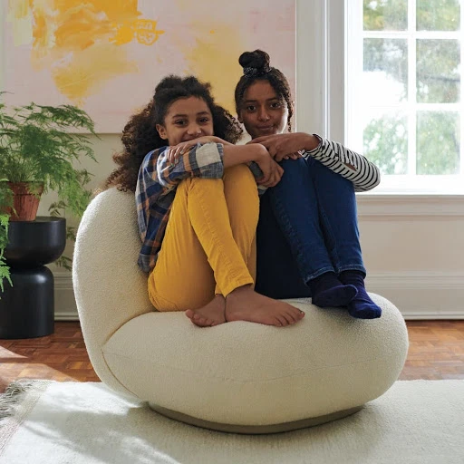 Two young girls sitting closely together on a large cream-colored, round chair indoors with natural light from a nearby window.