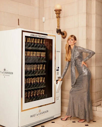 Elegant woman in a sequined gown poses beside a champagne vending machine inside a building with neutral tones and ornate lighting.