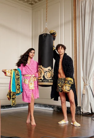 A man and woman in matching Versace robes stand next to a black Versace punching bag in an indoor setting, showcasing luxury loungewear.