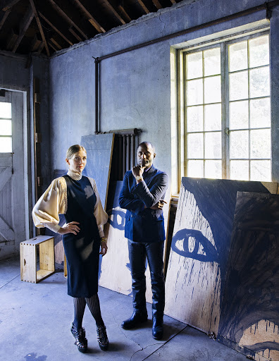 Two adults in an indoor space with art; woman in black dress and man in dark suit stand near canvases in a room with large windows.