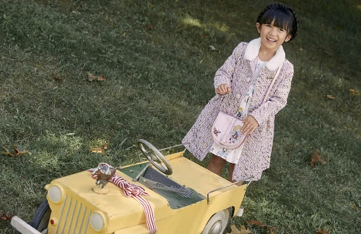 A happy young girl stands outside in a stylish coat next to a yellow toy car on a grassy lawn, smiling at the camera.