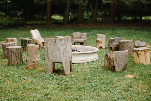 Outdoor gathering space features a circle of tree-stump chairs surrounding a white concrete fire pit on a green lawn beneath trees.