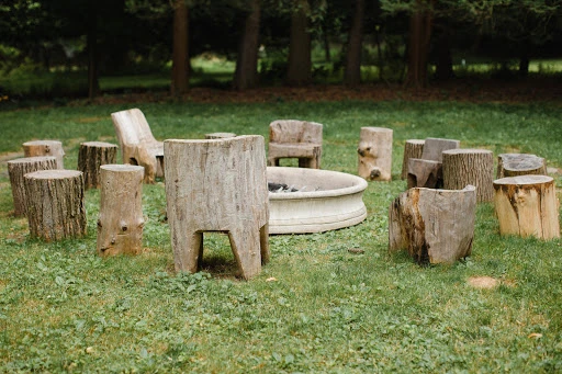 Outdoor gathering space features a circle of tree-stump chairs surrounding a white concrete fire pit on a green lawn beneath trees.