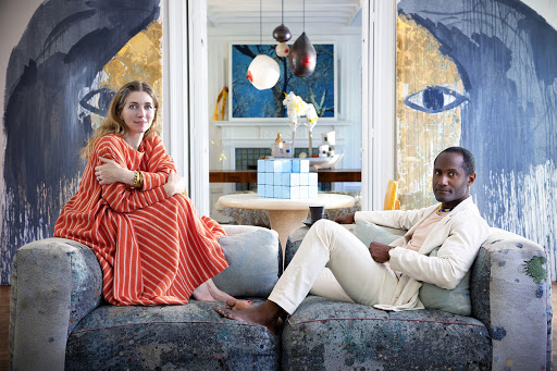 A woman in a striped orange dress and a man in light clothing seated on a large sofa in an artistic living room interior
