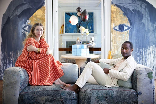 A woman in a striped orange dress and a man in light clothing seated on a large sofa in an artistic living room interior