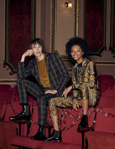 Two fashion models, an adult male and female, pose sitting in red theatre seats wearing a matching plaid suit and a patterned suit.