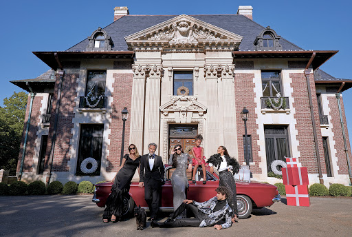 Elegant group poses by a red classic car in front of a mansion decorated for the holidays, featuring stylish formal wear and gift boxes.