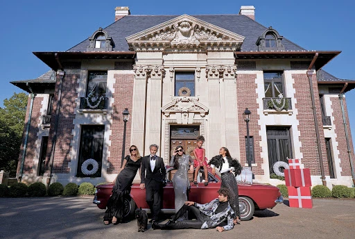 Elegant group poses by a red classic car in front of a mansion decorated for the holidays, featuring stylish formal wear and gift boxes.