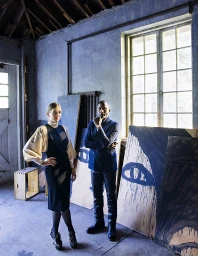 A man and woman standing inside an industrial studio space with large painted wooden panels leaning against the wall.