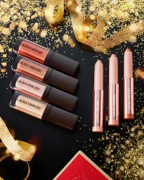 Four Laura Mercier cosmetic gloss tubes and three eyeshadow sticks on a black surface with gold glitter