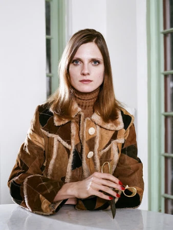 Portrait of a woman wearing a patchwork suede jacket and brown turtleneck, holding scissors in an interior space