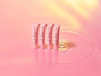 Four pink lip gloss tubes standing upright in shallow pink water with concentric ripples and soft warm lighting