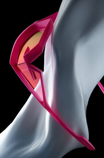 Close-up studio shot of angular, bright pink sunglasses with orange lenses positioned on a light grey flowing form against a black backdrop.