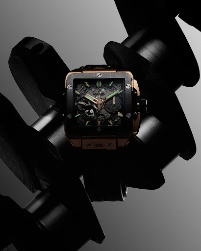 Elevated studio shot of a modern square-faced wristwatch with metallic details, positioned on a black mechanical engine component.