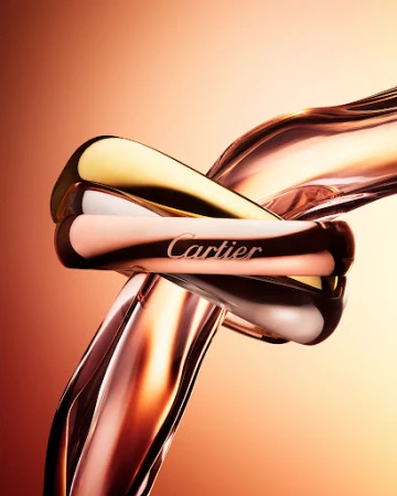 Close-up macro shot of interlocking gold, silver, and rose gold Cartier rings against a warm gradient background.