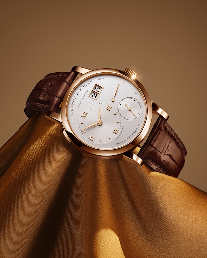 Luxury gold A. Lange & Söhne wristwatch with a brown leather strap resting on draped gold fabric in a studio with warm lighting.