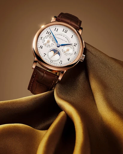 A. Lange & Söhne wristwatch floating on folded silk fabric, closeup studio shot, luxury timepiece, brown leather band, and gold case.
