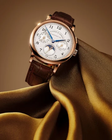 A. Lange & Söhne wristwatch floating on folded silk fabric, closeup studio shot, luxury timepiece, brown leather band, and gold case.