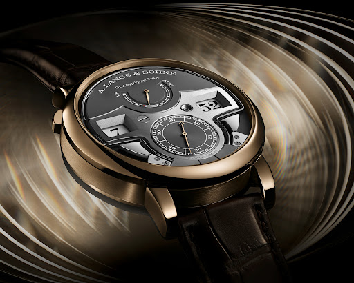 A close-up of a A. Lange & Söhne timepiece with a dark grey face, gold trim and dark leather strap, set against a blurred background.