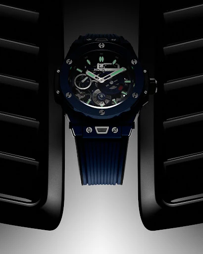 Close-up product shot of a dark blue luxury chronograph wristwatch with a ribbed rubber strap set against a dark architectural background.
