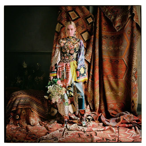 Fashion shot of a woman in a mixed-print dress standing in front of draped and layered carpets indoors with a bouquet of flowers.