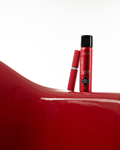 Cosmetic products from L'Oreal: a red tube of lipstick and a tall red can of Infallible 3-Second Setting Mist on a red glossy surface.