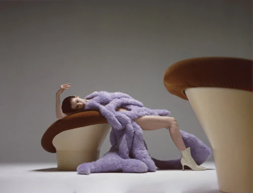 A woman in a lilac furry dress reclines on a tan chair next to another chair in a studio setting, creating an editorial fashion shot.