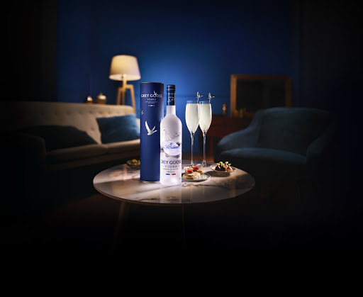 Grey Goose vodka bottle and filled champagne flutes are arranged on a marble table, with a lounge and lamp in the blurred background.