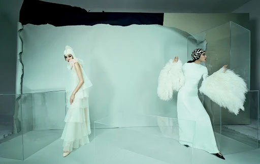 Two women pose in white dresses with head coverings in a staged setting with glass panels and a torn backdrop, fashion photography.