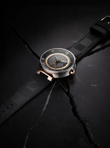 Luxury black wristwatch with gold accents displayed on dark marble with a textured rubber strap and a square patterned design on the band.
