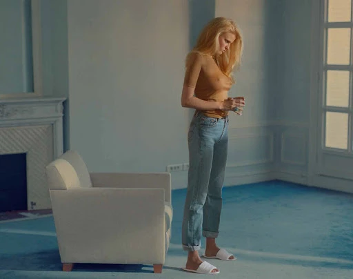 Full body portrait of an adult woman standing in a room with a chair and window, wearing jeans and a sheer top, holding a glass.