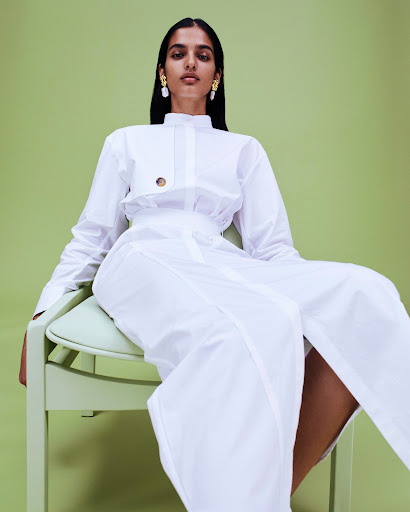 Woman in a white collared shirt and long white skirt seated on a pale green chair against a matching green background.