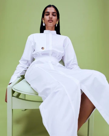 Woman in a white collared shirt and long white skirt seated on a pale green chair against a matching green background.