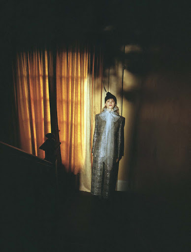 Adult female in a retro-inspired sparkly jumpsuit with a sheer ascot stands indoors, softly lit with a muted color palette.