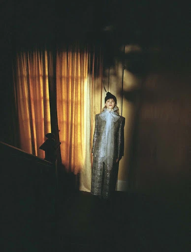 Adult female in a retro-inspired sparkly jumpsuit with a sheer ascot stands indoors, softly lit with a muted color palette.