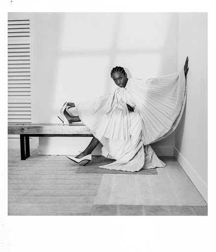 Black female model in a pleated white dress poses on a bench in a studio with white walls, minimalist style, and elegant fashion.