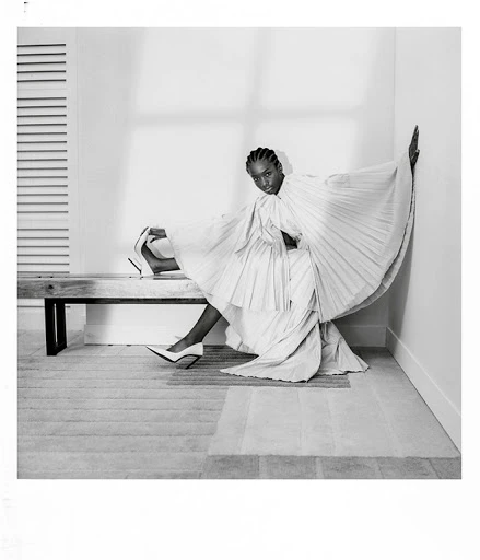 Black female model in a pleated white dress poses on a bench in a studio with white walls, minimalist style, and elegant fashion.