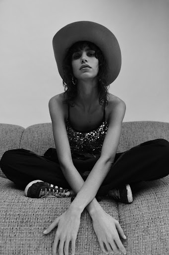 Monochrome portrait of a woman with a hat sitting on a couch. She has long, wavy hair and is wearing a sparkling tank top.