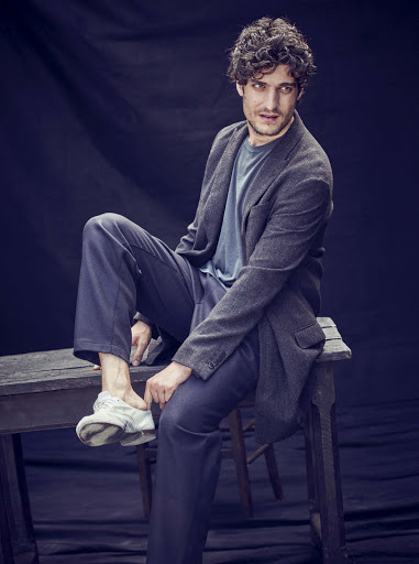 Man in gray blazer sitting on wooden stool against dark backdrop, wearing casual blue shirt, navy pants, and white sneakers in portrait.