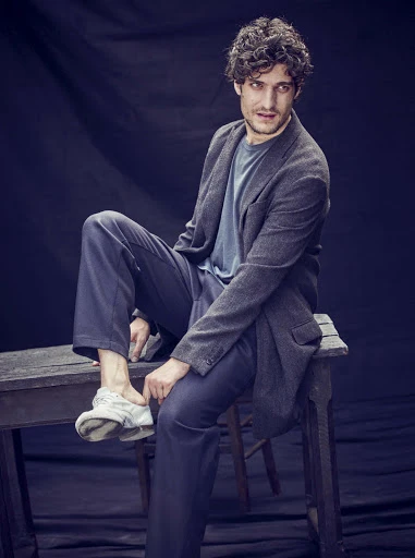 Man in gray blazer sitting on wooden stool against dark backdrop, wearing casual blue shirt, navy pants, and white sneakers in portrait.