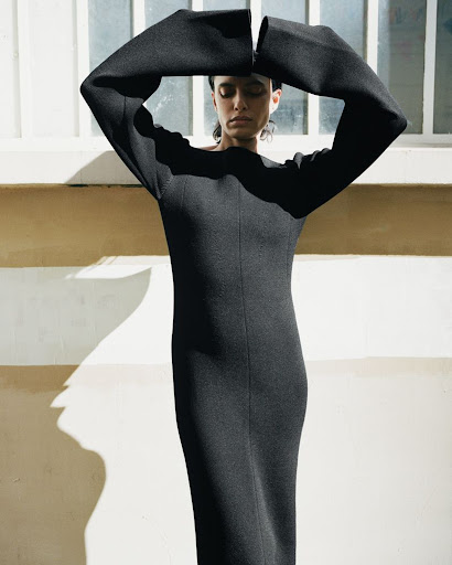 Model in a long black dress with arms raised overhead, set against an exterior wall with striped shadows.